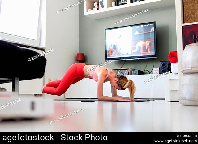 Attractive sporty woman working out at home, doing pilates exercise in front of television in her living room. Social distancing