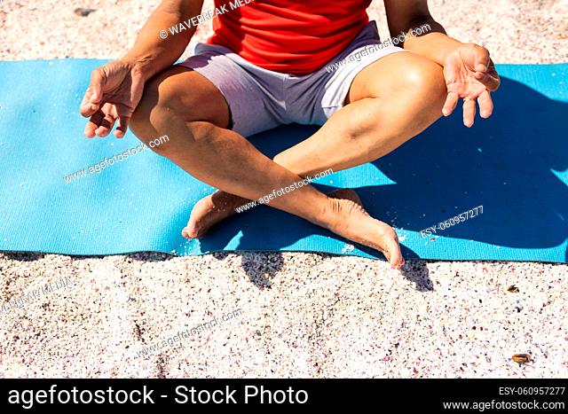 Low section of senior biracial man meditating in lotus position on yoga mat beach during sunny day