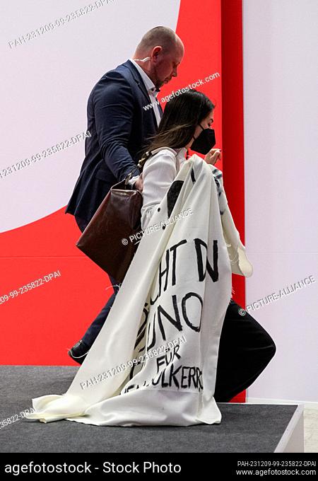09 December 2023, Berlin: A security guard leads a female activist off the stage during Chancellor Scholz's speech at the SPD's regular federal party conference...