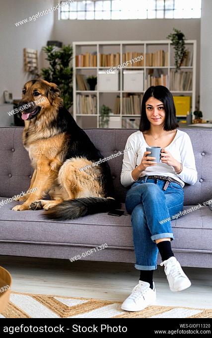 Young woman holding coffee cup while sitting with dog on sofa at home