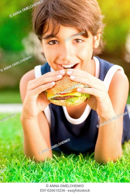 Portrait of a cute happy teen boy lying down on fresh green grass and with pleasure eating burger, tasty, but unhealthy food