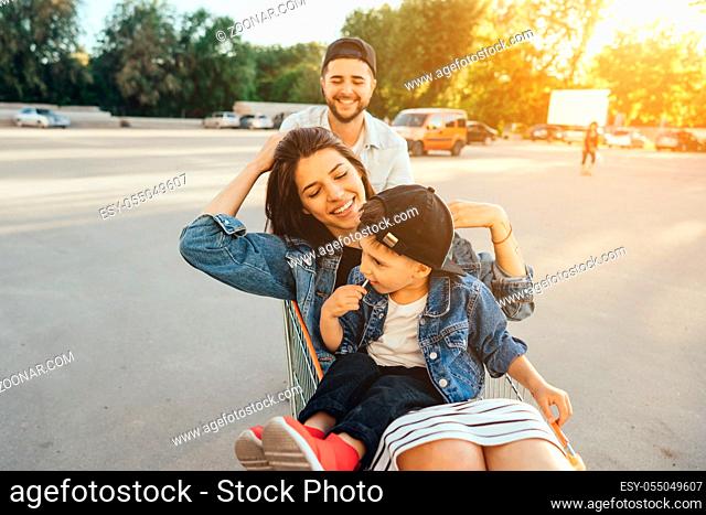 Young dad carries mom and son in a cart on the parking lot. The family is having fun