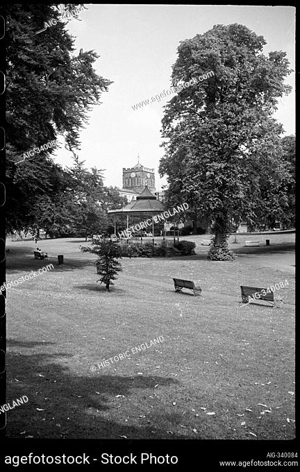General view of an early 20th century bandstand in a park, celled the Sele, with a partial view of Hexham Abbey with crossing tower in the background