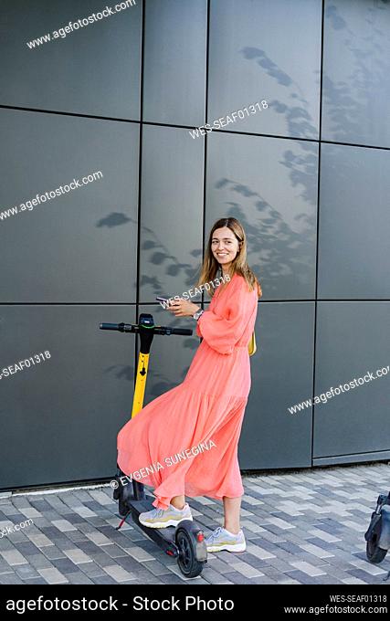Smiling woman holding mobile phone standing with electric push scooter on footpath