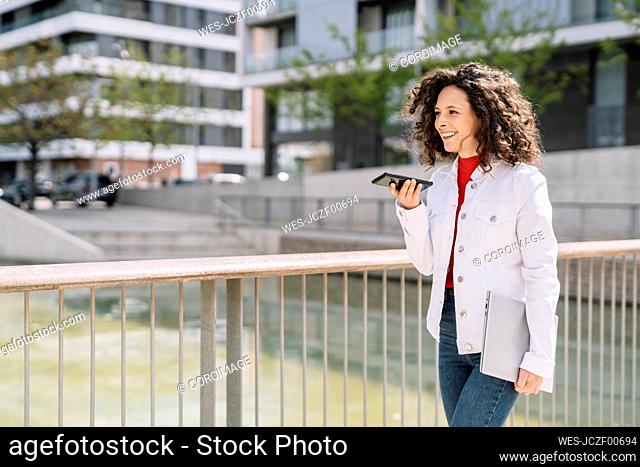 Cheerful woman sending voicemail through smart phone while walking by railing in city