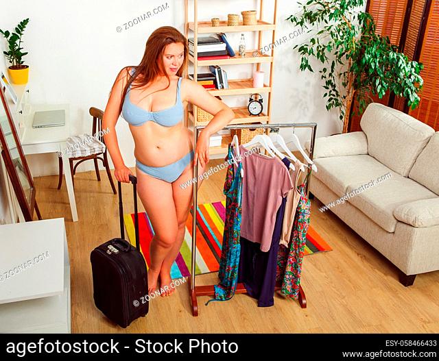 Portrait of beautiful woman trying different dresses at home. Pretty lady with red hair posing with luggage or suitcase