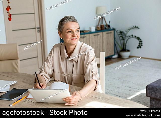 Thoughtful woman sitting at table at home with digital tablet and notebook
