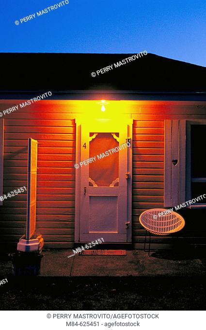 Illuminated roadside motel room entrance door and porch with chair at dawn