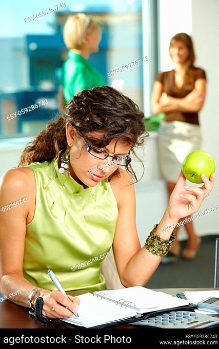 Young businesswoman sitting at desk in office, writing notes into personal organizer, holding apple