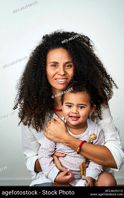 Smiling curly haired woman carrying daughter while sitting against gray background