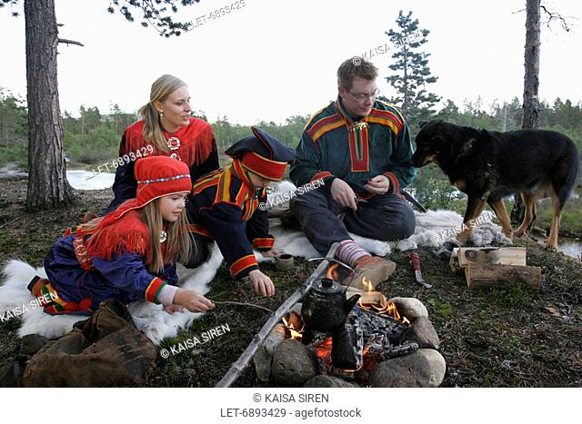 A Sami family dressed in the traditional Lapp dress and a Lapland reindeer dog are having a picnic by open fire in Inari, Finnish Lapland  Finland