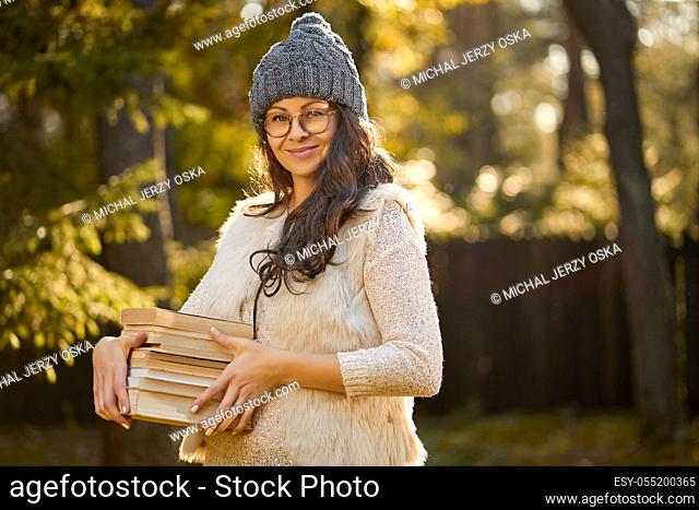 beautiful woman in a cap and glasses is holding a pile of books on the background of autumn forest