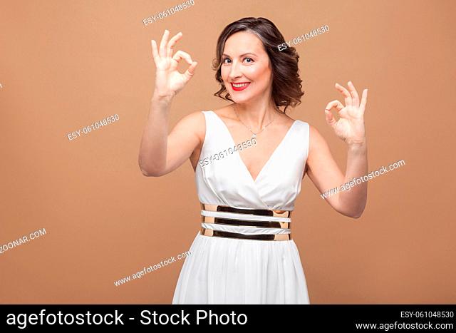 Toothy smiled woman in white dress showing ok sign. Emotional expressing woman in white dress, red lips and dark curly hairstyle
