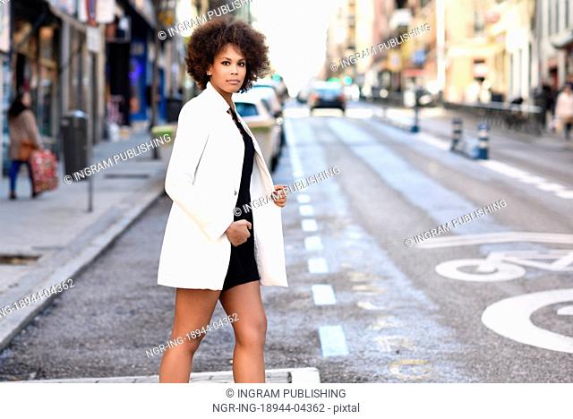 Young black woman with afro hairstyle walking on a crosswalk in an urban street. Mixed girl wearing white jacket and black dress with city background