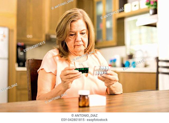 Good looking senior adult woman looking at her pills' expiration date sitting in the kitchen