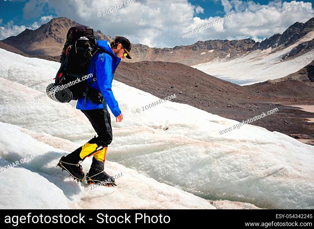 A mountaineer with a backpack walks in crampons walking along a dusty glacier with sidewalks in the hands between cracks in the mountain