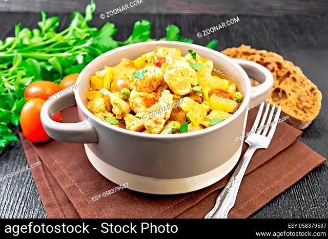 Stew of chicken breast, tomatoes, stalked celery, carrots, green peas and onions in a tureen on a napkin against dark wooden board