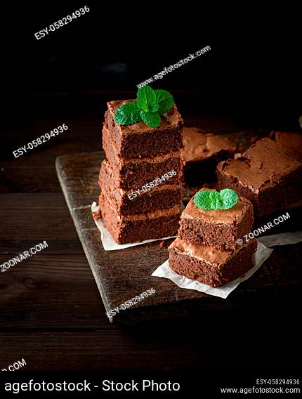 stack of square pieces of baked brown brownie pie on a wooden board, on the top is a green mint leaf