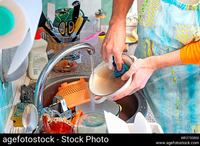 A man is washing dishes in kitchen. He is using a sponge and dishwashing product. Hot water flows from the tap