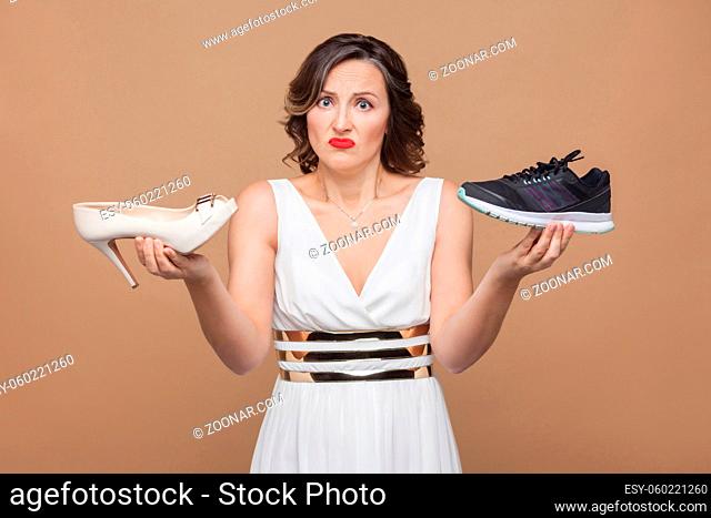 Doubtful thinking business woman in white dress standing, holding elegant high heels and comfortable sneakers and don't know which one choose