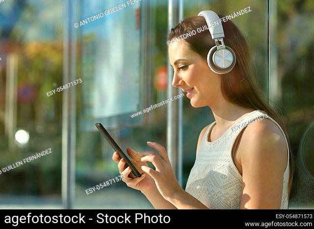 Side view portrait of a commuter listening to music with headphones and smart phone in a bus stop