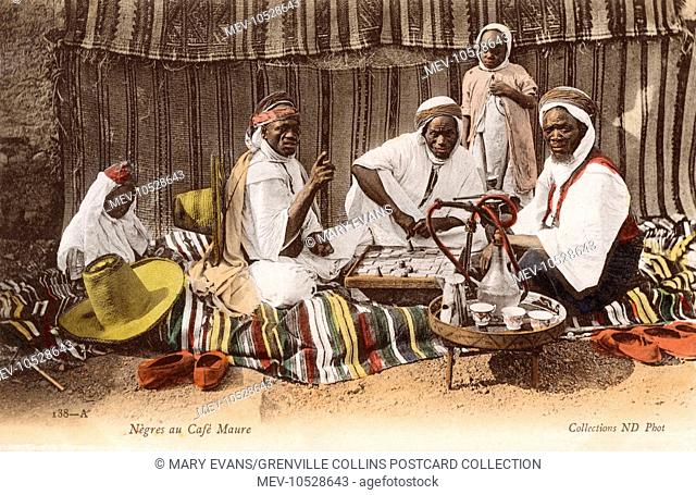 Black African men taking tea and a smoke of a hookah pipe at a drink at a Moorish Cafe - Algeria. Two of the men are playing a board game