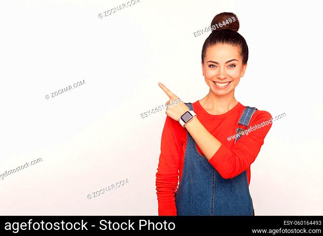 Attention to advertising. Portrait of happy stylish pretty girl with hair bun in denim overalls pointing aside to copy space, blank wall for commercial text