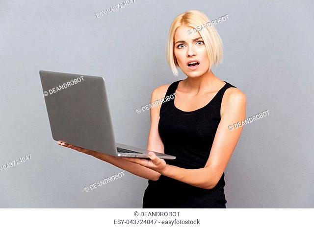 Amazed unhappy young woman using laptop over gray background