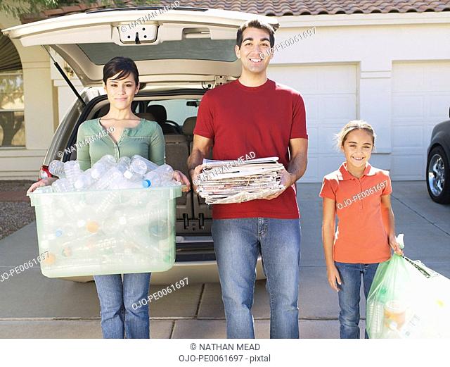 Man and woman with young girl outdoors standing in driveway holding recyclable materials and smiling