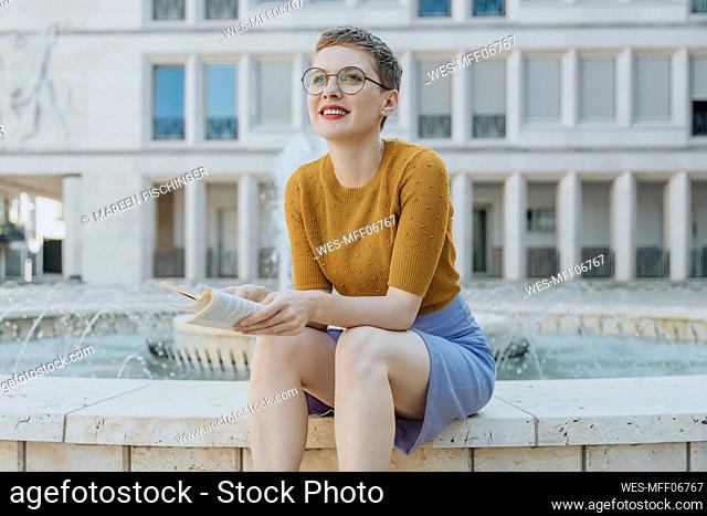 Mid adult woman looking away with book sitting against fountain