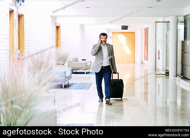 Male entrepreneur on phone pulling wheeled luggage in hotel corridor