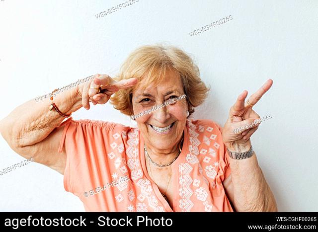 Happy senior woman gesturing peace sign in front of wall