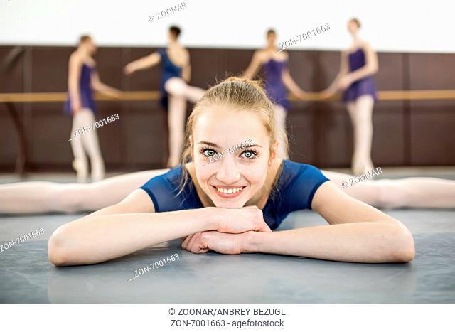 ballerina sitting on the floor with his face to the camera in the splits and dance class dancers practicing on the background