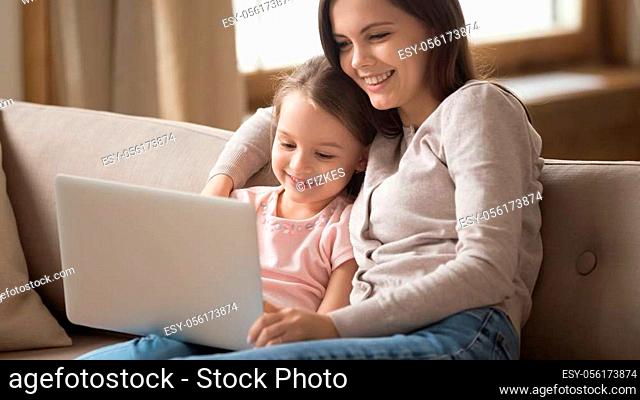 Happy young mother and little daughter using laptop together, hugging, sitting on couch at home, smiling mum and adorable preschool girl looking at laptop...