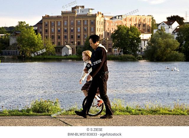 woman with child on a an unicycle at Ruhr river in Hattingen with Birschel mill in background, Germany, North Rhine-Westphalia, Ruhr Area, Hattingen