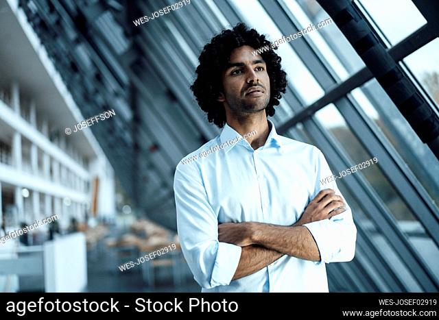 Thoughtful young businessman standing with arms crossed while looking away at workplace