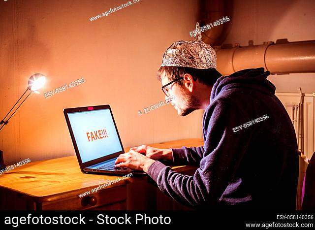 Young man with aluminum cap is sitting in the dark basement in front of a laptop. Conspiracy theory concept
