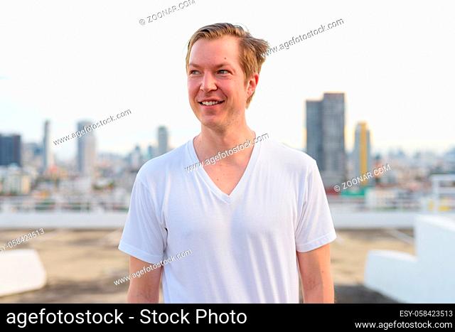 Portrait of young handsome Scandinavian man with blond hair against view of the city at rooftop of building outdoors