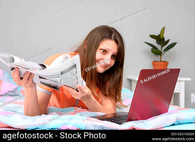 A girl with newspapers in her hands lies in bed and looks at the laptop screen in search of work