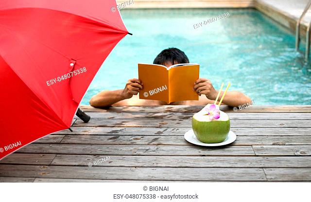 Man relaxing reading a book and having coconut juice with flower by the swimming pool