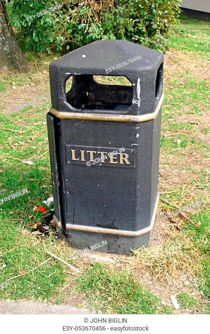 Worn black plastic litter bin with some litter beside it. Surrounded by mown grass and shrubs behind