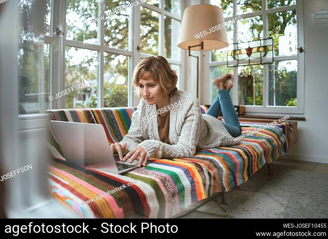 Smiling woman using laptop lying on sofa at home