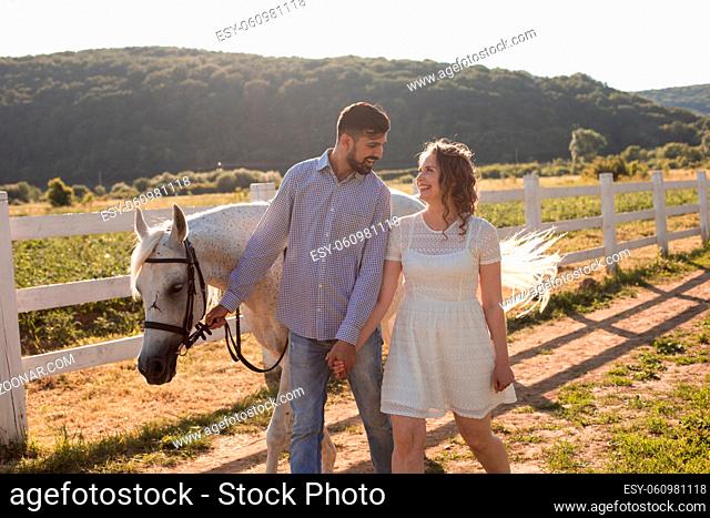 Couple walk at the ranch. The young bearded man leads a white horse