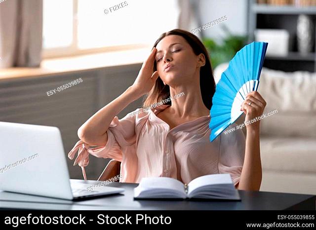 Overheated woman waving fan, sitting at desk with laptop at home, stressed young female suffering from heating at home, feeling discomfort