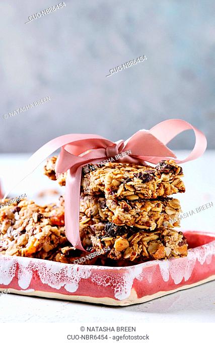 Homemade energy oats granola bars with dried fruits and nuts whole and broken with pink ribbon in ceramic plates on grey table. Healthy snack