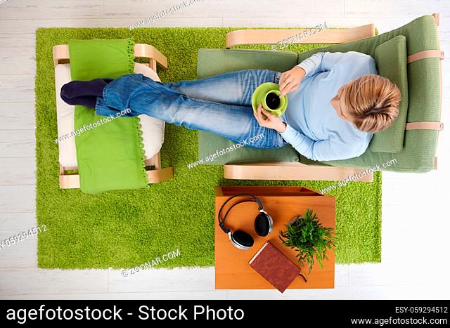 Woman having coffee sitting in armchair with legs crossed on footboard in high angle view