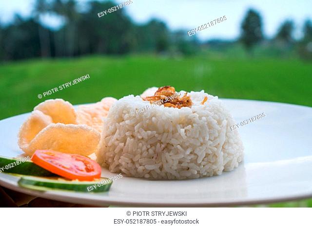 Steamed rice with prawn crackers and vegetables on white plate over blurred rice fields background