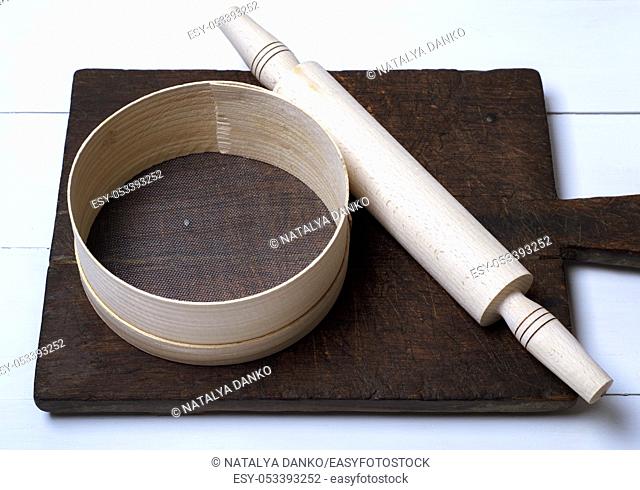 brown wooden kitchen cutting board, rolling pin and sieve on a white wooden table, top view