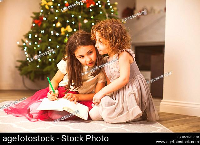 Two adorable girl friends sharing their secret dreams about Christmas presents, writing letter to Santa. Sisters sitting close on a carpet in warm living room...