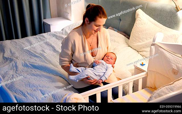 Young mother sitting next to baby crib with her newborn child at night in bedroom
